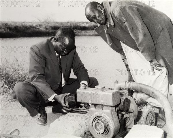 Reservoir water pump. A interested by-stander observes as a man in a suit inspects a reservoir pump designed to supply homes with water for domestic use. Kenya, circa 1965. Kenya, Eastern Africa, Africa.