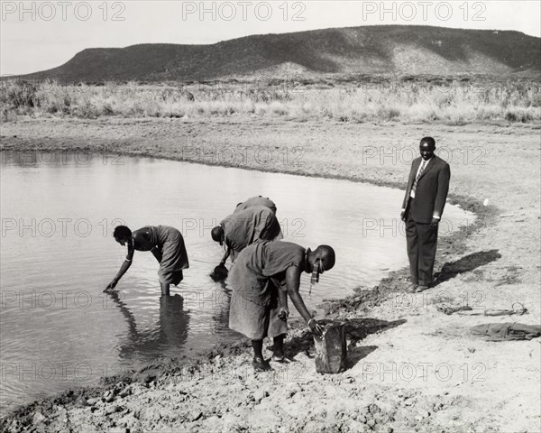Collecting water in a dry zone. A man in a suit observes as women in traditional dress collect water from a lake on the site of a newly constructed dam built to conserve water for farmers in a dry zone. Kenya, circa 1965. Kenya, Eastern Africa, Africa.