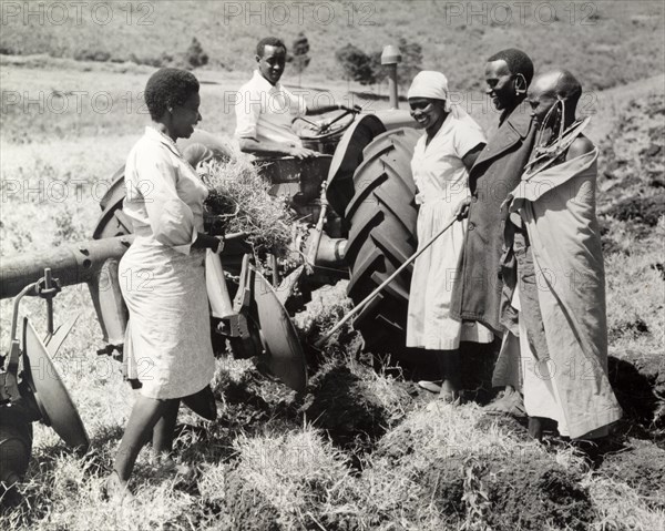 A communal ploughing team. A team of African farm workers pause to chat around a tractor pulling a large plough. The pristine work clothes worn by three of the workers contrast with the traditional dress and jewellery of the other two. Kenya, circa 1965. Kenya, Eastern Africa, Africa.