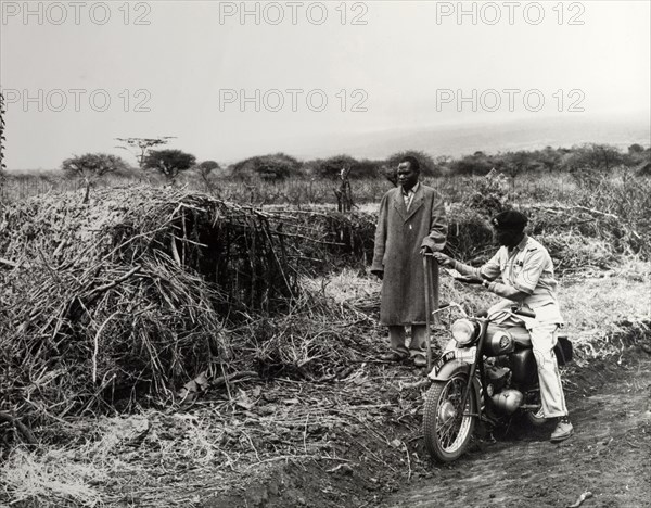 Agricultural officer. A uniformed agricultural instructor offers advice to an African farmer as he passes on his motorcycle. Notebook in hand, he points to a structure made from sticks by the side of the track. Kenya, circa 1965. Kenya, Eastern Africa, Africa.