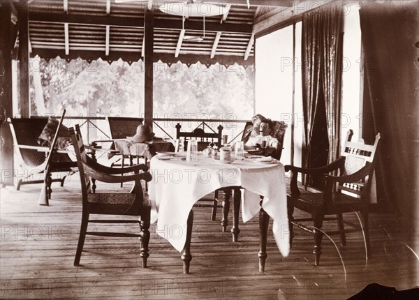 Colonial veranda. A European man smokes a cigar as he reclines in a chair on a colonial veranda. His shotgun rests upright against an armchair: a dining table set for two dominates the foreground. Possibly Burma (Myanmar), South East Asia, circa 1910., South East Asia, Asia.
