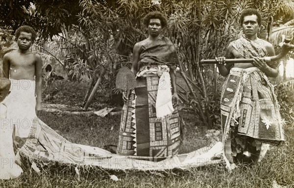Fijian ceremonial costume. A Fijian man and woman pose for the camera dressed in traditional ceremonial attire. The woman holds up a basketwork fan, the man a curved stick. Fiji, circa 1920. Fiji, Pacific Ocean, Oceania.
