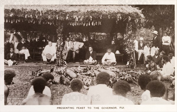 Feast for the Governor, Fiji. An audience of Fijian locals are gathered around a thatched hut, decorated in honour of the Governor of Fiji who is being presented with a feast. Fiji, circa 1920. Fiji, Pacific Ocean, Oceania.