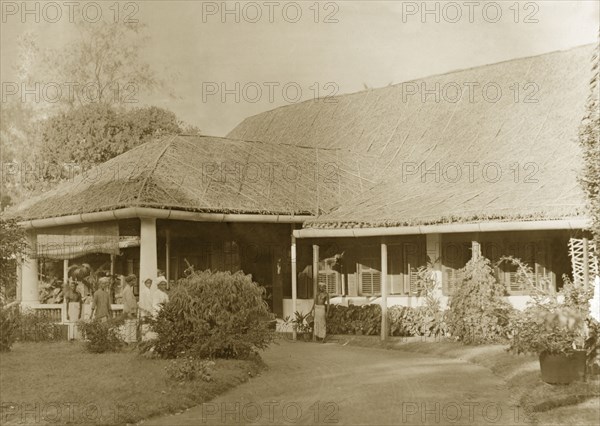 Colonial bungalow, Burma (Myanmar). Several servants stand on the driveway at the entrance to a large colonial bungalow with a thatched roof and shuttered windows. Probably Burma (Myanmar), circa 1946. Burma (Myanmar), South East Asia, Asia.