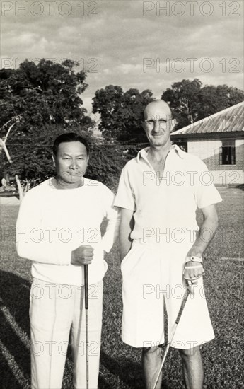 Golf partners, Rangoon. R.B. Groves, a Captain of the Rangoon Golf Club, poses for a portrait mid-game alongside an unidentified Burmese man. Both men are dressed in their sporting whites, golf clubs in hand. Rangoon (Yangon), Burma (Myanmar), circa 1952. Yangon, Yangon, Burma (Myanmar), South East Asia, Asia.