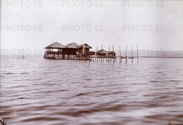 Lake dwellings in Burma (Myanmar). Two wooden dwellings on stilts project from the calm waters of a lake. Burma (Myanmar), circa 1910. Mohnyin, Kachin, Burma (Myanmar), South East Asia, Asia.