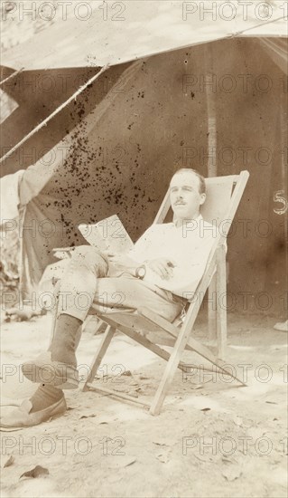 Relaxing in camp. A European man reads a book whilst reclining in a deckchair under the canopy of a tent. Probably Burma (Myanmar), circa 1905. Burma (Myanmar), South East Asia, Asia.