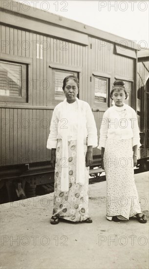 Burmese women at a railway station. Portrait of two Burmese women standing on a railway platform in front of a stationary, second-class train carriage. They wear patterned 'longyis' with blouses and sandals. Burma (Myanmar), circa 1940. Burma (Myanmar), South East Asia, Asia.
