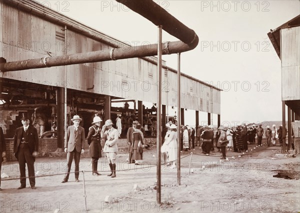 Tour of an industrial factory. A group of smartly dressed European men and women take a tour of an industrial factory, pausing beside a shed filled with large, cylindrical containers and a network of piping. Several turbaned Sikh men are also present. Probably India, circa 1940. India, Southern Asia, Asia.