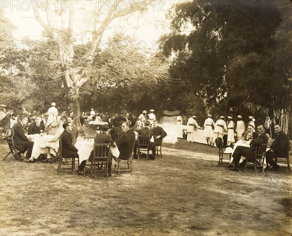Guests at a garden party. Small groups of European men and women sit around dining tables in the garden of a colonial residence, waiting to be served by Indian servants who dish out food from a long buffet table. India, circa 1925. India, Southern Asia, Asia.