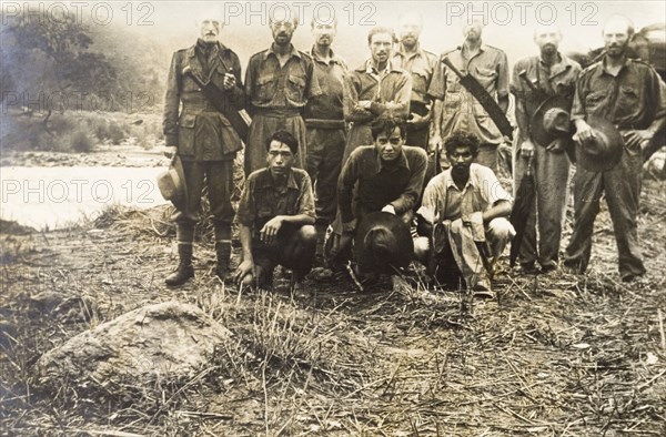 A trekking party in the jungle. Eight European men pose for a group portrait beside a river with their Asian assistants. Some carry machetes, suggesting the group were on a trek through dense jungle, perhaps a surveying expedition. India, June 1942. India, Southern Asia, Asia.
