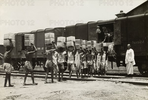 Queuing up to load a freight train. A team of workers carry boxes on their heads and form a queue, as they load cargo onto a freight car under the direction of an overseer. India, circa 1942. India, Southern Asia, Asia.