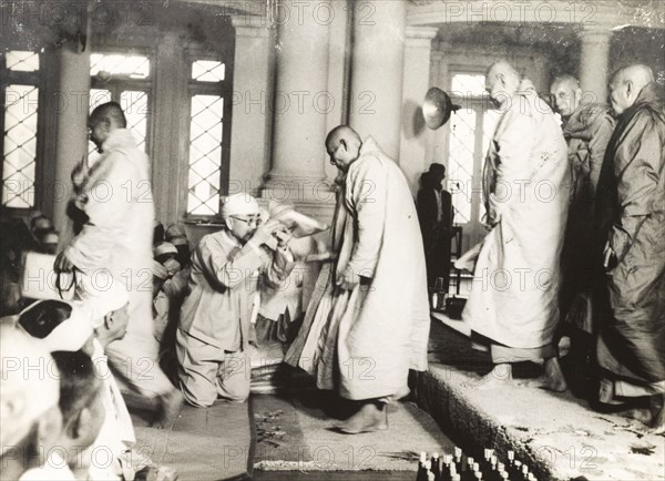 Buddhist monks enter a temple. A kneeling Burmese man hands out padded kneelers to a number of robed Buddhist monks as they file into a temple. Burma (Myanmar), circa 1943. Burma (Myanmar), South East Asia, Asia.