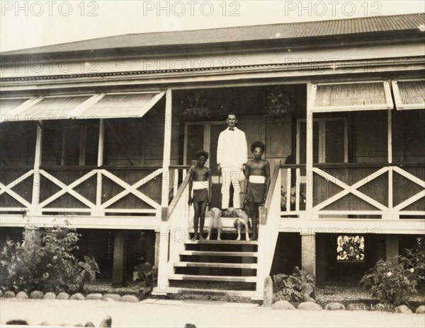 Clifford Francis and his domestic servants. A European District Officer, Clifford Francis, poses for the camera on the steps of his colonial-style bungalow, accompanied by two Solomon Islander domestic servants and two dogs. Gizo, Solomon Islands, 1919. Gizo, West (Soloman Islands), Solomon Islands, Pacific Ocean, Oceania.