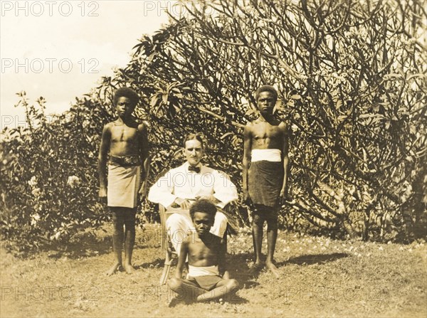 Clifford Francis poses with his servants. Outdoors portrait of Clifford Francis, a European District Officer, posing in a chair beside three of his Solomon Islander domestic servants. Gizo, Solomon Islands, 1919. Gizo, West (Soloman Islands), Solomon Islands, Pacific Ocean, Oceania.