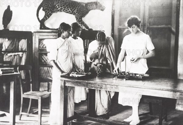 Botany lesson at a Women's Christian College. Four female students (three Indian and one European) study botany at the Women's Christian College in Madras. They examine specimens through a microscope in a classroom equipped with cabinets full of books and a stuffed leopard. Madras, Madras Presidency (Chennai, Tamil Nadu), India, 1918. Chennai, Tamil Nadu, India, Southern Asia, Asia.