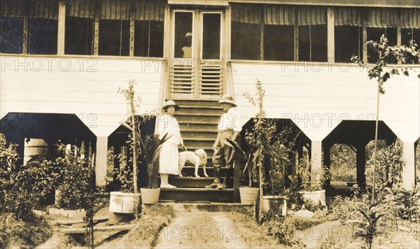European couple on steps of colonial house. A European couple, identified as Mr and Mrs Johnson, pose on the steps of a large, colonial stilt house with their pet dog, whilst a servant watches from the doorway. British Guiana (Guyana), 1927. Guyana, South America, South America .