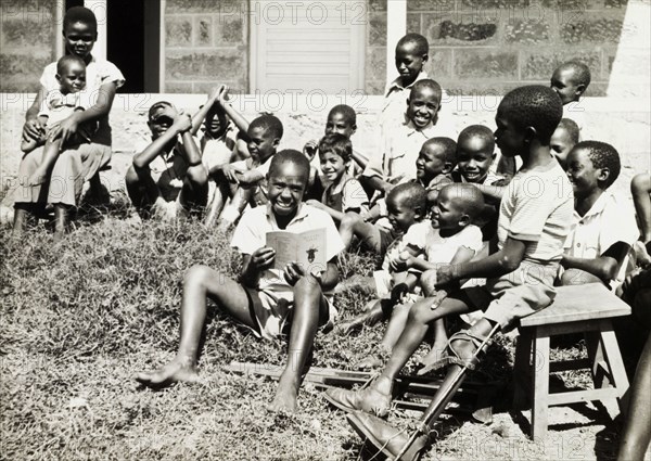Children at a charity in Nairobi. An older boy reads a story to a group of younger children on a patch of grass outdoors. A disabled boy to his left sits on a stool with his leg bound in a splint. An original caption comments that this boy 'had appealed to us to be able to wear (his crutches) and so walk again', after his father had pawned his walking aids twice, and suggests that these children were receiving help from a European charity of some sort. Nairobi, Kenya, March 1955. Nairobi, Nairobi Area, Kenya, Eastern Africa, Africa.