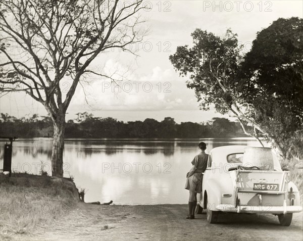 Man gazing over picturesque lake. A solitary man leans against a loaded car parked by the side of a picturesque lake, as he gazes out over the calm water. Probably Northern Rhodesia (Zambia), circa 1950. Zambia, Southern Africa, Africa.