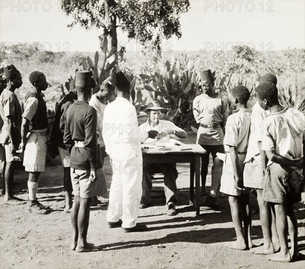 African workers collecting their pay. Young African men in Western-style clothing form a queue at a table where a European man sits smoking and handling envelopes, possibly handing out pay packets to his employees. Lusaka, Northern Rhodesia (Zambia), circa 1950. Lusaka, Lusaka, Zambia, Southern Africa, Africa.