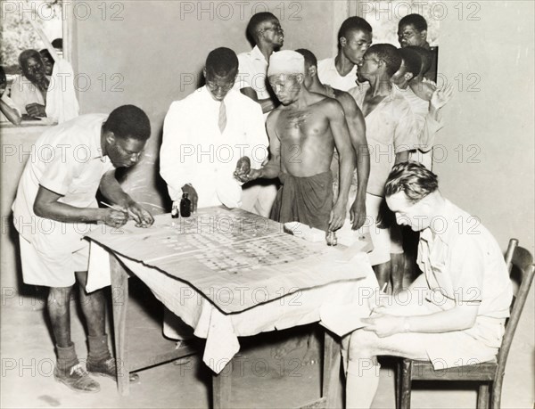 Blood samples at village dispensary. A group of African men queue at a dispensary, where two African medics are taking blood samples. A European doctor sits writing notes next to a table, on which the blood samples are laid out on slides. Probably Northern Rhodesia (Zambia), circa 1950. Zambia, Southern Africa, Africa.