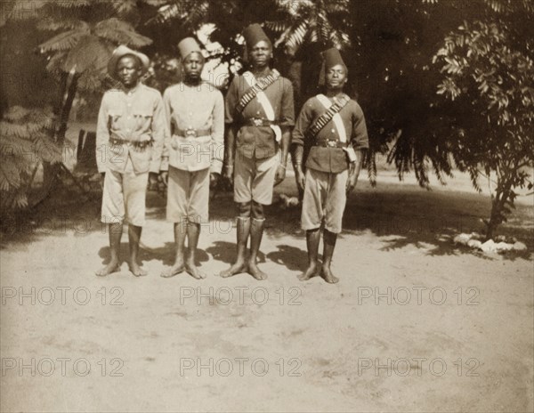 African Policemen. Outdoors portrait of four uniformed African policemen. They stand barefoot in a row, two of the officers wearing bandoliers and fez hats. Probably Northern Rhodesia (Zambia), circa 1950. Zambia, Southern Africa, Africa.