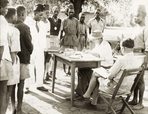 Performing administrative duties. A European man sits at a table writing in a book, with two piles of envelopes positioned in front of him. He is surrounded by a queue of African men in Western-style clothing, possibly handing out pay packets to his employees. Lusaka, Northern Rhodesia (Zambia), circa 1950. Lusaka, Lusaka, Zambia, Southern Africa, Africa.
