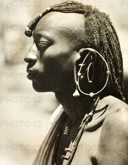 African man with a distinctive earring. Profile shot of the head and shoulders of a young African man. Naked from the waist up, he wears a plaited hairstyle, necklaces and a large, distinctive earring. Probably Northern Rhodesia (Zambia), circa 1950. Zambia, Southern Africa, Africa.