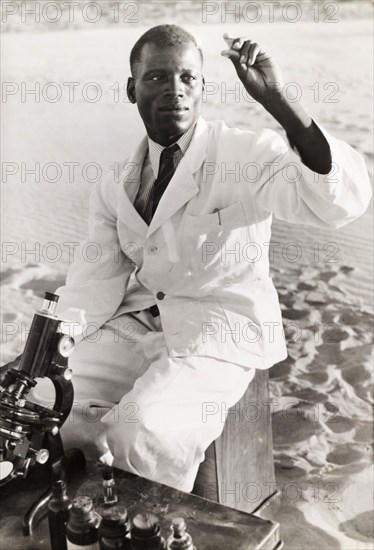 An African medic inspects a blood sample. An African medic holds up and inspects a blood sample on a slide, as he sits in front of a microscope at a makeshift desk, whilst undertaking field work. Probably Northern Rhodesia (Zambia), circa 1950. Zambia, Southern Africa, Africa.