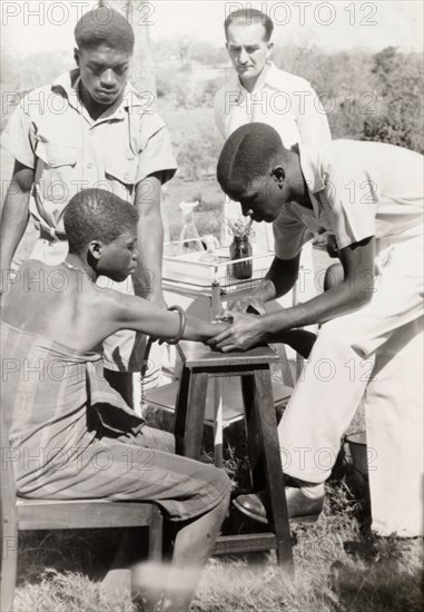 An African medic injects a patient. An African medic gives an injection to a female patient, while a European doctor stands by a medical trolley and observes. Probably Northern Rhodesia (Zambia), circa 1950. Zambia, Southern Africa, Africa.