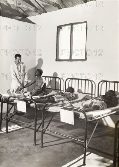 A European doctor examines African patients. A European doctor examines a patient as he sits on a hospital bed in a ward containing two other male patients. Probably Northern Rhodesia (Zambia), circa 1950. Zambia, Southern Africa, Africa.