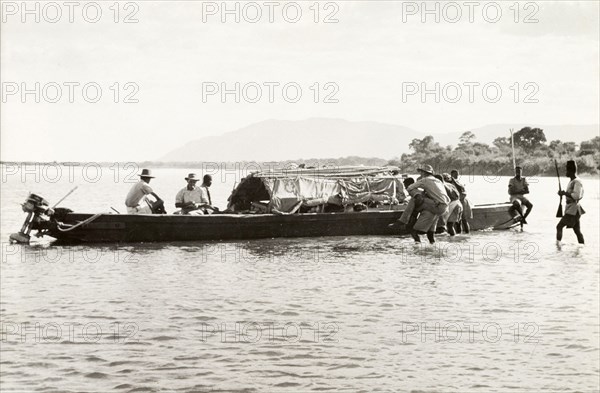 Boarding the District Commissioner's River boat. A European man is carried through shallow water to the District Commissioner's motor boat by an African crew member. Two European men are already onboard the boat, sitting at the bow for the vessel, ready to depart. Probably Northern Rhodesia (Zambia), circa 1950. Zambia, Southern Africa, Africa.