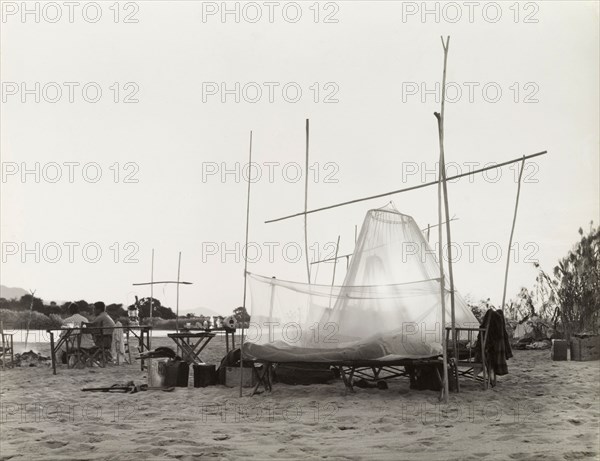 Camping on the banks of the Zambezi River. The camp site of a District Commissioner on the banks of the Zambezi River, where tents consisting of mosquito nets are strung up on a framework of wooden poles. Two men sit by a camp fire, watching dawn break over the river. Northern Rhodesia (Zambia), circa 1950. Zambia, Southern Africa, Africa.