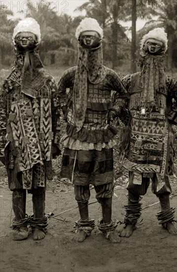 Juju practitioners. Three juju dancers in costume wear wooden masks on top of their heads, ornate patterned clothing and anklets. Possibly Badagry, Nigeria, circa 1925. Nigeria, Western Africa, Africa.