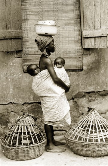 Yoruba woman with 'pickins'. A barefooted Yoruba woman wearing a headscarf carries two babies strapped close to her body, one on her back and one on her belly. She stands sideways on between two baskets used for transporting chickens and balances a large bowl on her head. Nigeria, circa 1930. Nigeria, Western Africa, Africa.