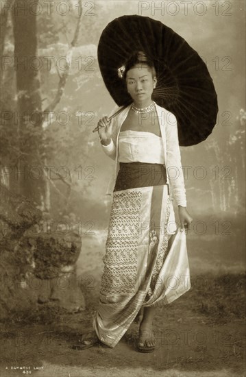Portrait of a Burmese lady. A studio portrait of a young Burmese woman, elegantly posed with a parasol and finely dressed in a bodice, jacket and a patterned silk 'longyi' or wraparound skirt. Her hair is worn up with a decorative hair ornament and her outfit is completed with jewelled earrings and a matching necklace. Burma, circa 1885. Burma (Myanmar), South East Asia, Asia.