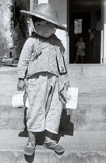 Refugee from China. A refugee boy from China stands on steps infront of an open doorway with an identity card and towel pinned to his shirt. He wears a straw hat and holds a tin cup in one hand. Hong Kong, People's Republic of China, 1963. Hong Kong, Hong Kong, China, People's Republic of, Eastern Asia, Asia.