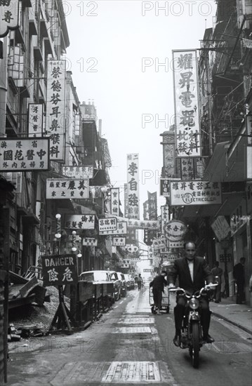 Hong Kong side street. A lively side street in the city centre crowded with signs written in Chinese and English. Hong Kong, People's Republic of China, 1963. Hong Kong, Hong Kong, China, People's Republic of, Eastern Asia, Asia.