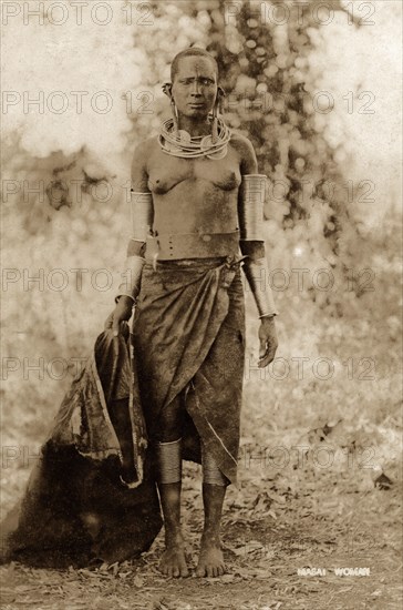 Maasai woman. Full-length portrait of a semi-naked Maasai woman wearing metal necklaces, ear, arm and leg ornaments. She has a bark cloth wrapped around her waist and carries an animal skin sack. Kenya, circa 1930. Kenya, Eastern Africa, Africa.