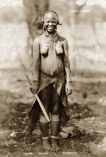 Kikuyu woman smiling. Full-length portrait of a smiling Kikuyu woman wearing neck, ear, arm and leg ornaments. She has a bark cloth wrapped around her waist and holds a knife for clearing bush, a reference to the Kikuyu's expertise as cultivators. Kenya, circa 1930. Kenya, Eastern Africa, Africa.
