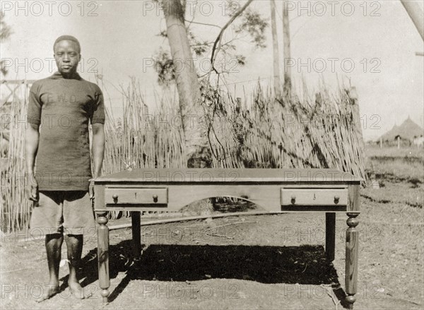 Cabinetmaker with desk. A Public Works Department employee, identified in the the caption as 'Witaba Wa Sitamani', stands beside a desk with side drawers and turned legs that he has made. He wears a government uniform that displays the initials 'PWD' on the chest. British East Africa (Kenya), March 1916. Kenya, Eastern Africa, Africa.