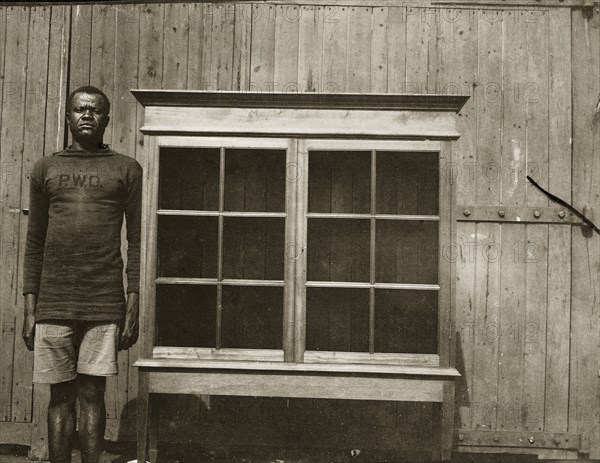 Cabinet for Empire Exhibition. An employee of the Public Works Department stands beside a cabinet with glass doors that he has made for display at the Empire Exhibition taking place at Wembley, London in 1924. He wears a government uniform that displays the initials 'PWD' on the chest. Nakuru, Kenya, 1924. Nakuru, Rift Valley, Kenya, Eastern Africa, Africa.
