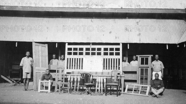 Cabinetmakers from the Public Works Department. Local employees of the Kenyan Public Works Department pose beside an array of doors, window frames, chairs and other wooden furniture constructed by them under the supervision of Charles Bungey, a training officer with the PWD. Nakuru, Kenya, 1924. Nakuru, Rift Valley, Kenya, Eastern Africa, Africa.