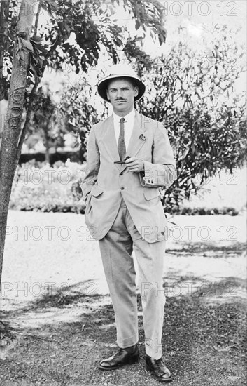 European man with solatopi. Informal portrait of a European man wearing a lightweight suit and solatopi, an Indian sun hat made from the pith of the sola plant. He holds a pipe in his left hand and stands next to a tree marked with the number 68. Kenya, circa 1920. Kenya, Eastern Africa, Africa.