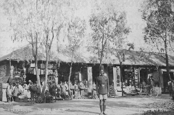 Indian market. A policeman pictured at an Indian market in front of a range of single-storey shops crowded with people and goods. British East Africa (Kenya), 1912. Kenya, Eastern Africa, Africa.