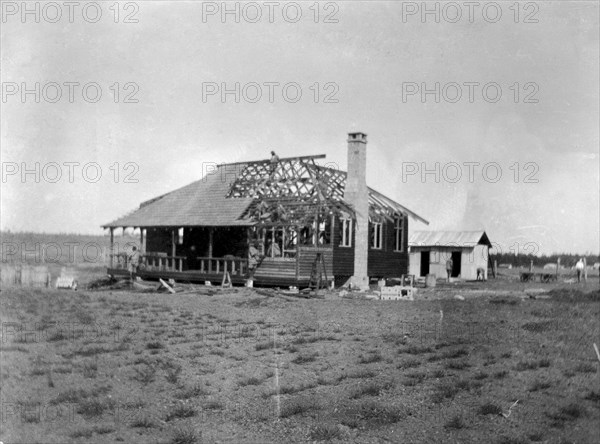 Building a house. This is the first in a series of four photographs showing the rapid erection of a timber-framed European-style house. The photograph shows the house from the front, with a stone chimney rising up the side. The house was built for Charles Bungey, a training officer with the Kenyan Public Works Department, by the local apprentices of his department. Nakuru, Kenya, March 1922. Nakuru, Rift Valley, Kenya, Eastern Africa, Africa.