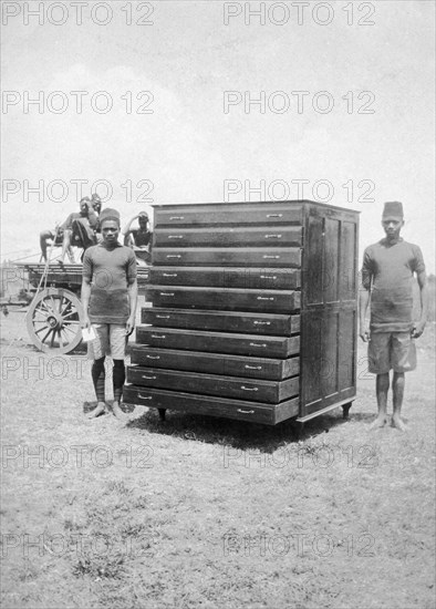 African cabinetmakers. Two young African men stand either side of a large map cabinet they have made as employees of the Public Works Department. Both men wear government uniforms and are identified in the caption as 'Makongoro wa Chancoro (sic) na Juma Wasila'. British East Africa (Kenya), 1914. Kenya, Eastern Africa, Africa.