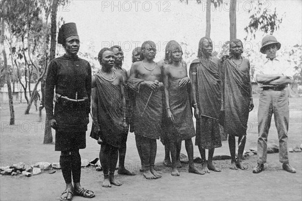 Raw recruits. A European officer and an African soldier stand either side of a small group of traditionally dressed African men, all new recruits for the King's African Rifles. British East Africa (Kenya), circa 1916. Kenya, Eastern Africa, Africa.