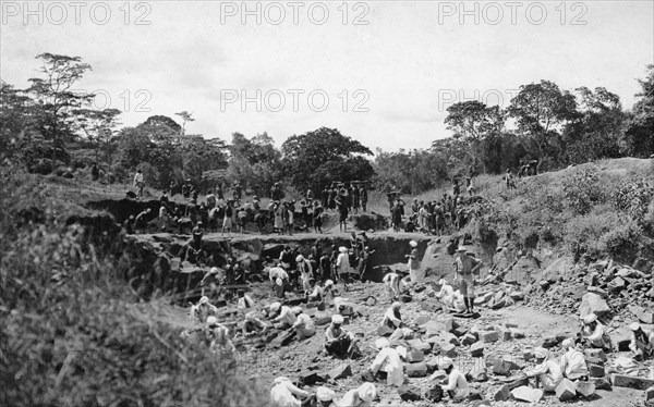 Stone quarry. A European man stands in the centre of a shallow quarry surrounded by African and Indian workers cutting and carrying stones. Nairobi, British East Africa (Kenya), 1913. Nairobi, Nairobi Area, Kenya, Eastern Africa, Africa.