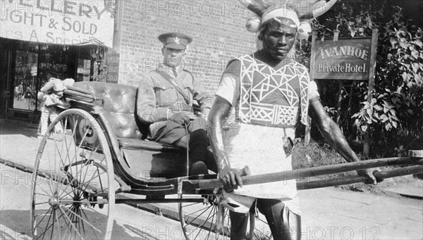 Rickshaw puller in Durban. An African rickshaw puller wearing a horned headdress and a decorated bib, is pictured with his client, a British army officer, outside the Ivanhoe Private Hotel. Durban, Natal (KwaZulu Natal), South Africa, circa 1915. Durban, KwaZulu Natal, South Africa, Southern Africa, Africa.
